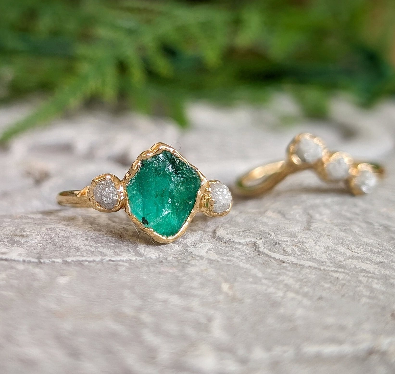 Raw emerald and rough diamond Chevron wedding ring set on a textured surface, featuring unique natural stones and 18k gold.
