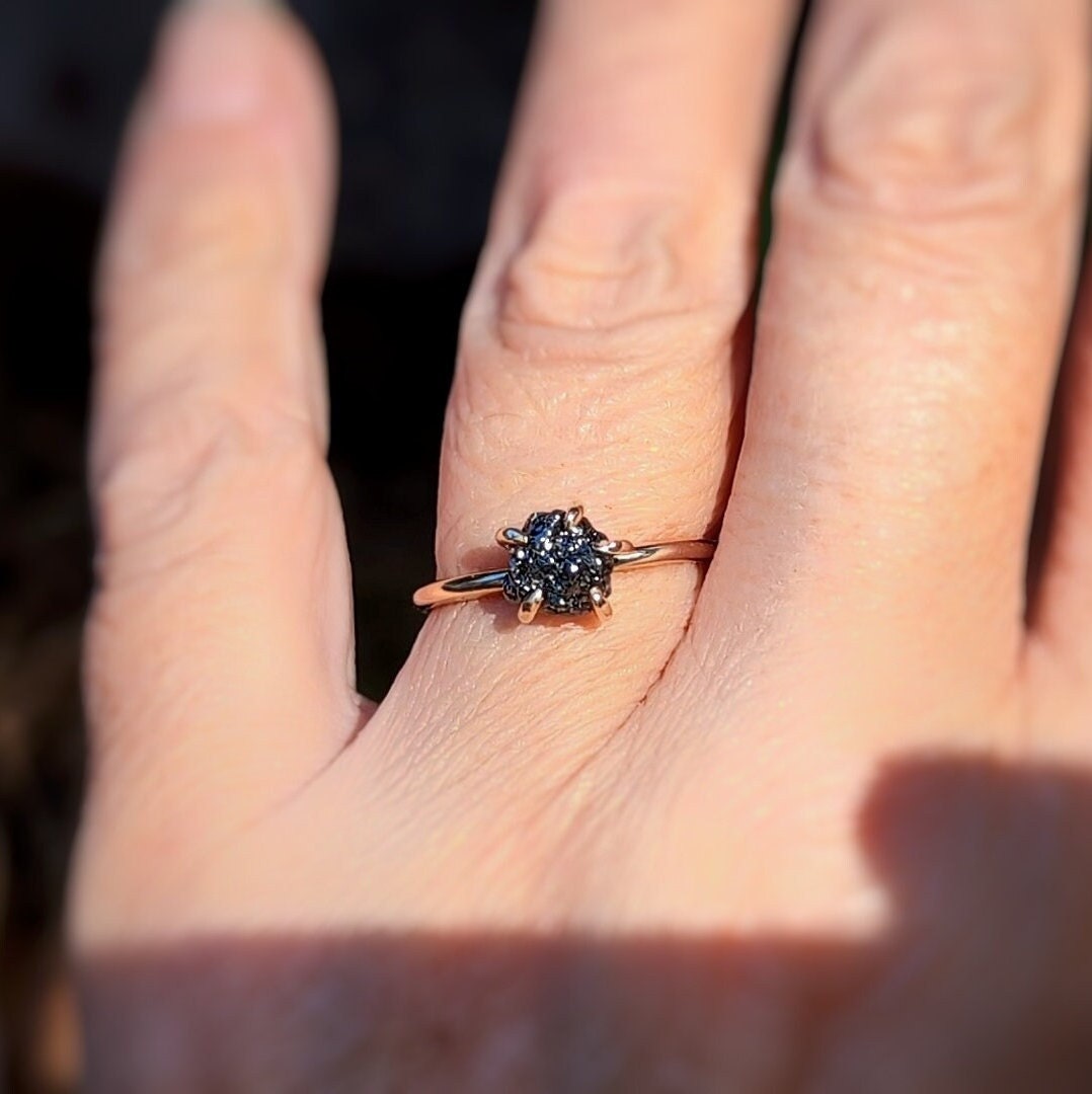 Raw black diamond engagement ring on hand, featuring 14k gold band and 7-8mm uncut black diamond.