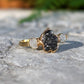 Raw uncut Black Diamond engagement ring on a stone surface, featuring two smaller raw diamonds and a 14k Gold band.