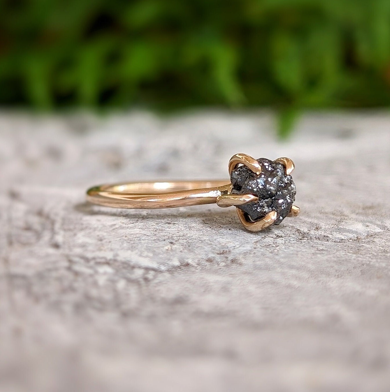 Raw black diamond engagement ring on a stone surface with a 14k gold band, showcasing a unique uncut design.