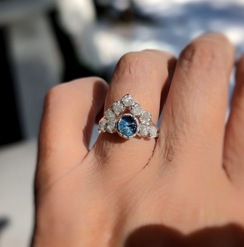 Raw Montana Sapphire and diamond bridal ring set showcased on hand, featuring teal blue sapphire and conflict-free diamonds.
