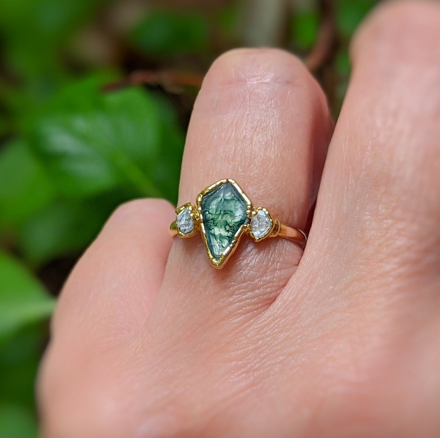 Kite-shaped Moss Agate ring with Herkimer diamonds, symbolizing love and elegance, displayed on a hand amidst greenery.