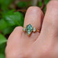 Kite-shaped Moss Agate ring with Herkimer diamonds, symbolizing love and elegance, displayed on a hand amidst greenery.