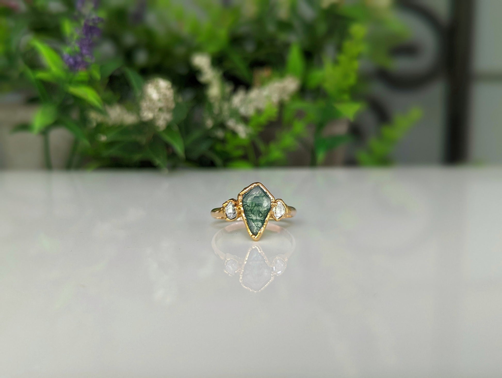 Kite-shaped Moss Agate and Herkimer diamond engagement ring on a reflective surface with greenery in the background.