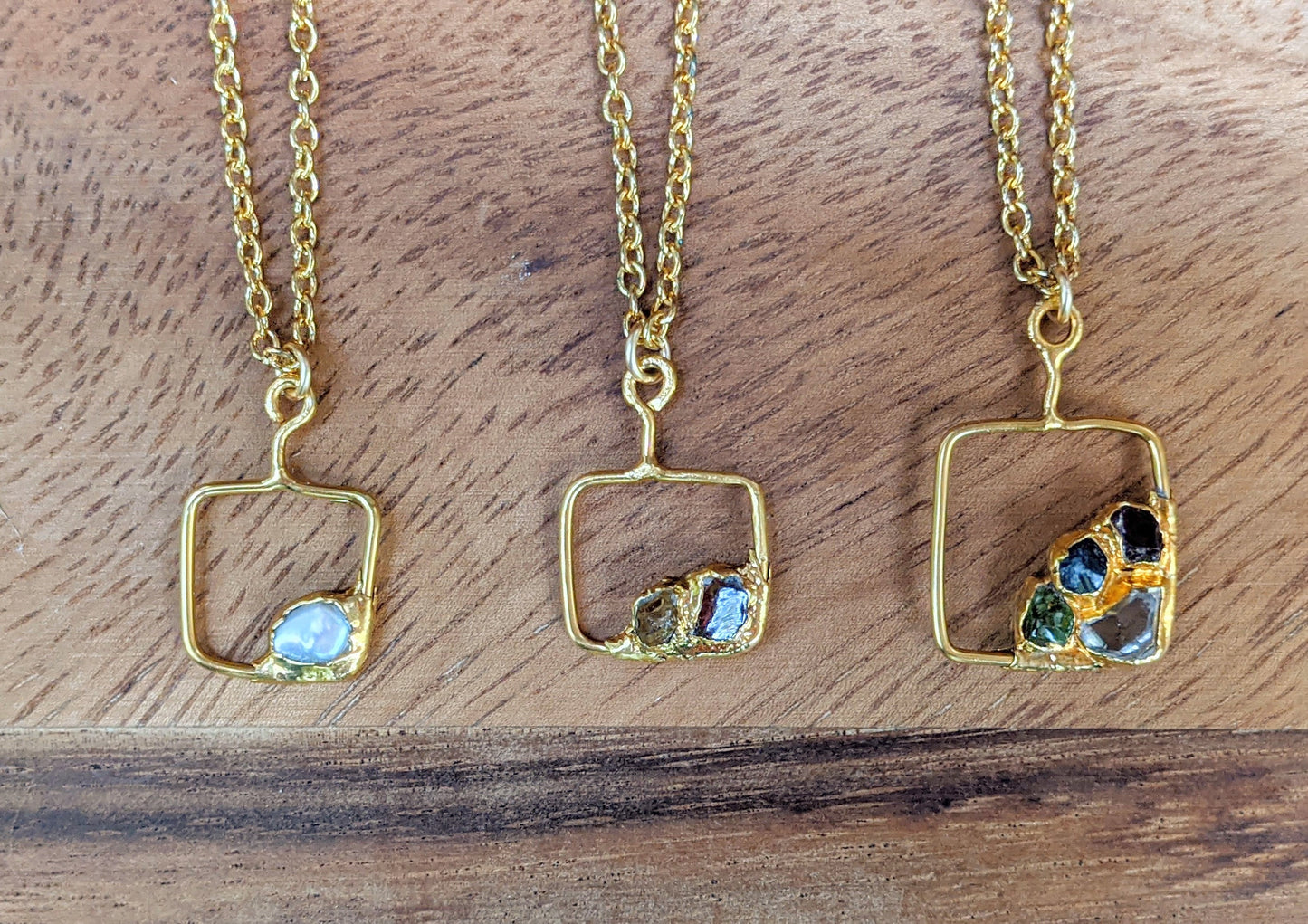 Three personalized gold necklaces with unique square pendants and raw gemstones on a wooden background.
