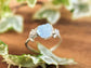 Raw aquamarine and diamond twig engagement ring on a wooden surface surrounded by green leaves.