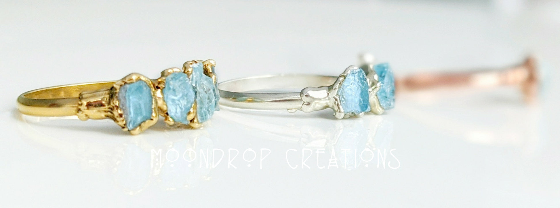 Three raw Aquamarine rings on display: gold, silver, and rose gold bands featuring natural gemstones.