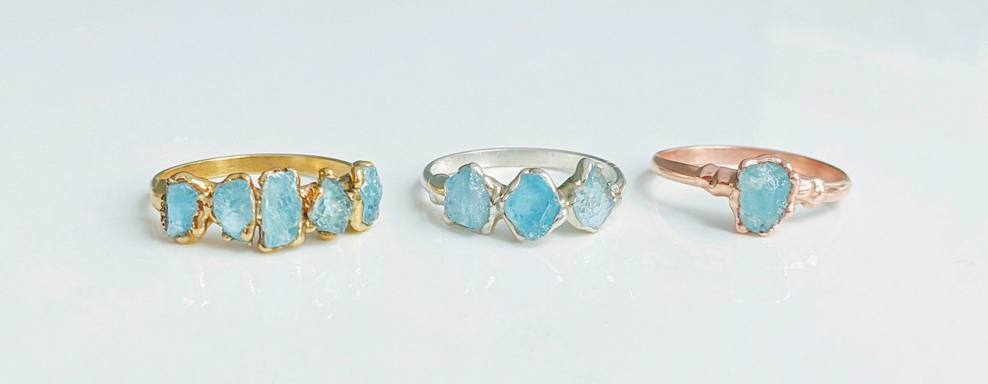 Three raw Aquamarine rings on display: gold, silver, and rose gold bands with natural stones, perfect March birthstone jewelry.