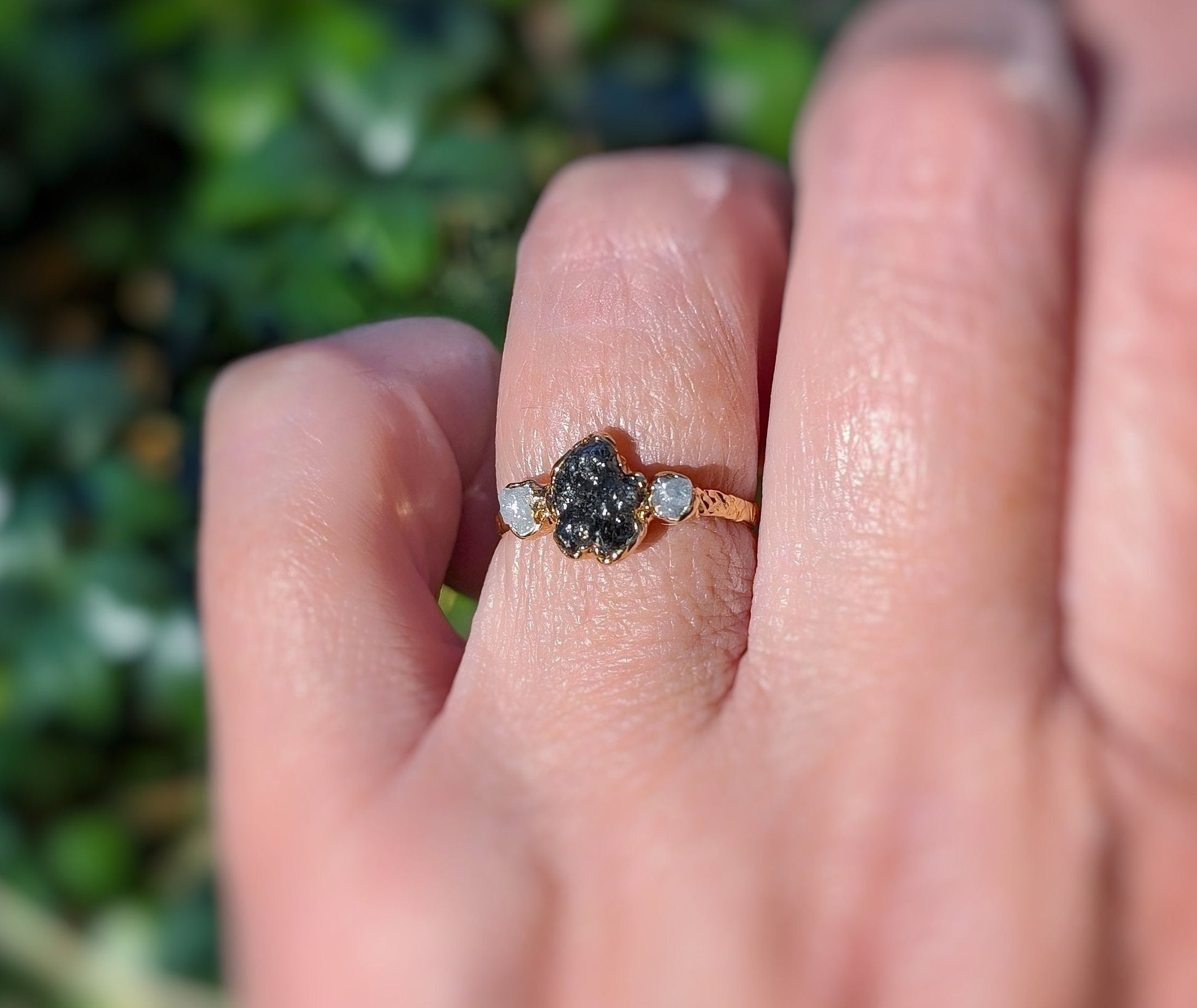 Raw black diamond engagement ring with rough diamond centerpiece and smaller diamonds on band, hand model against green background.
