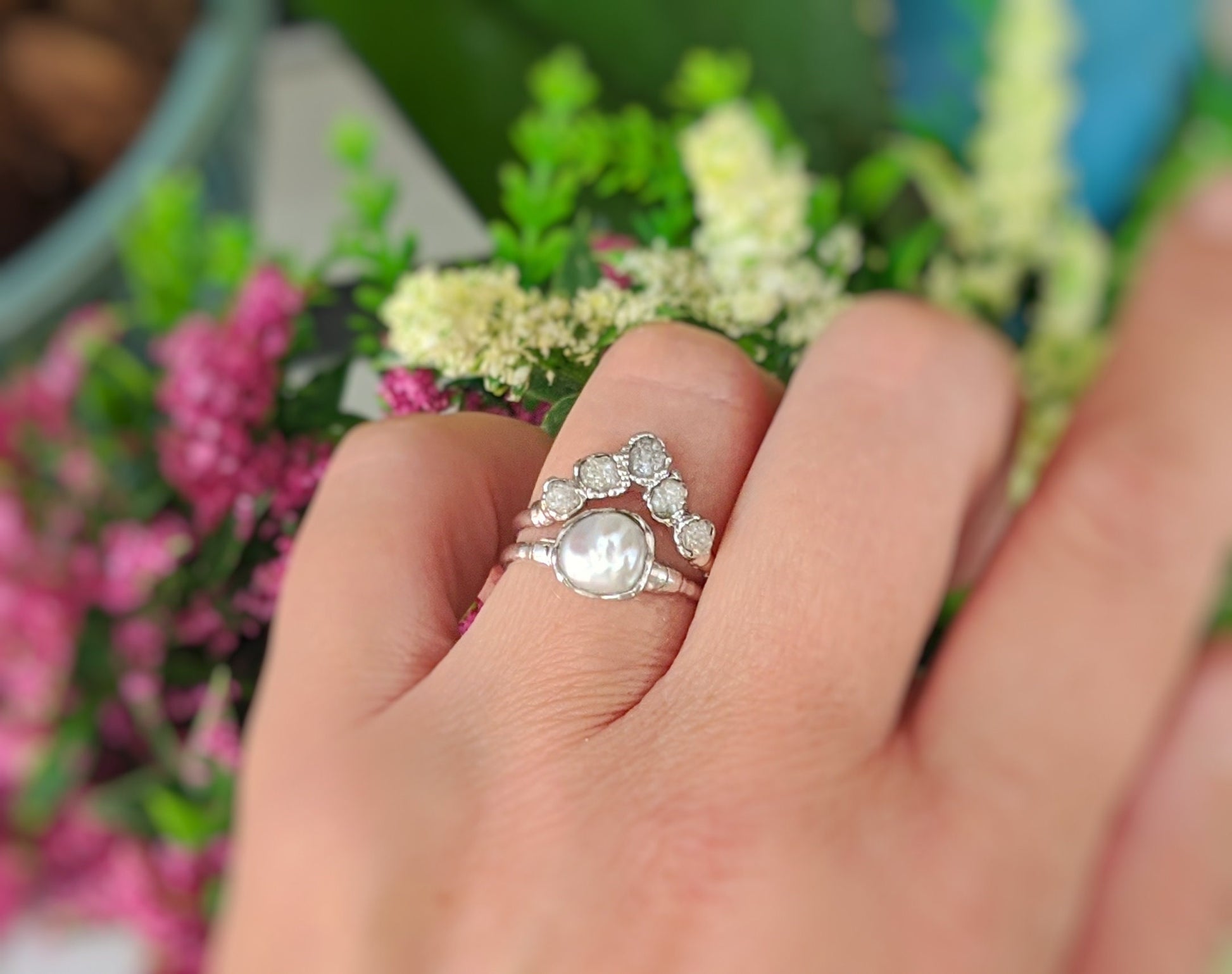 Elegant raw diamond and Keshi pearl Chevron wedding ring set worn on a hand, surrounded by vibrant flowers.