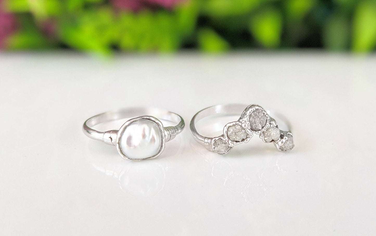 Raw diamond and Keshi pearl Chevron wedding ring set on a reflective surface with greenery in the background.