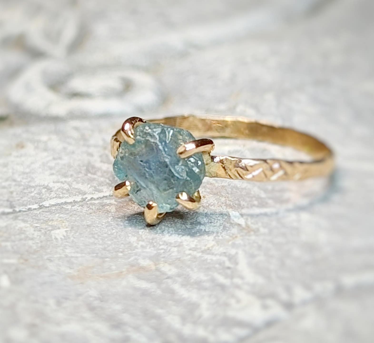 Teal-blue raw Montana Sapphire engagement ring with textured gold band on a stone surface.
