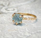 Teal-blue raw Montana Sapphire engagement ring with textured gold band on a stone surface.