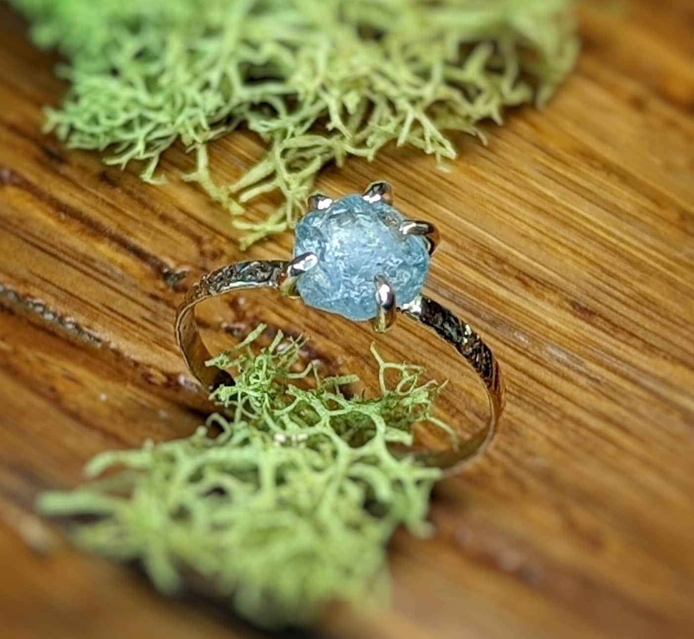 Teal-blue raw Montana Sapphire engagement ring on a rustic wooden surface with green moss.