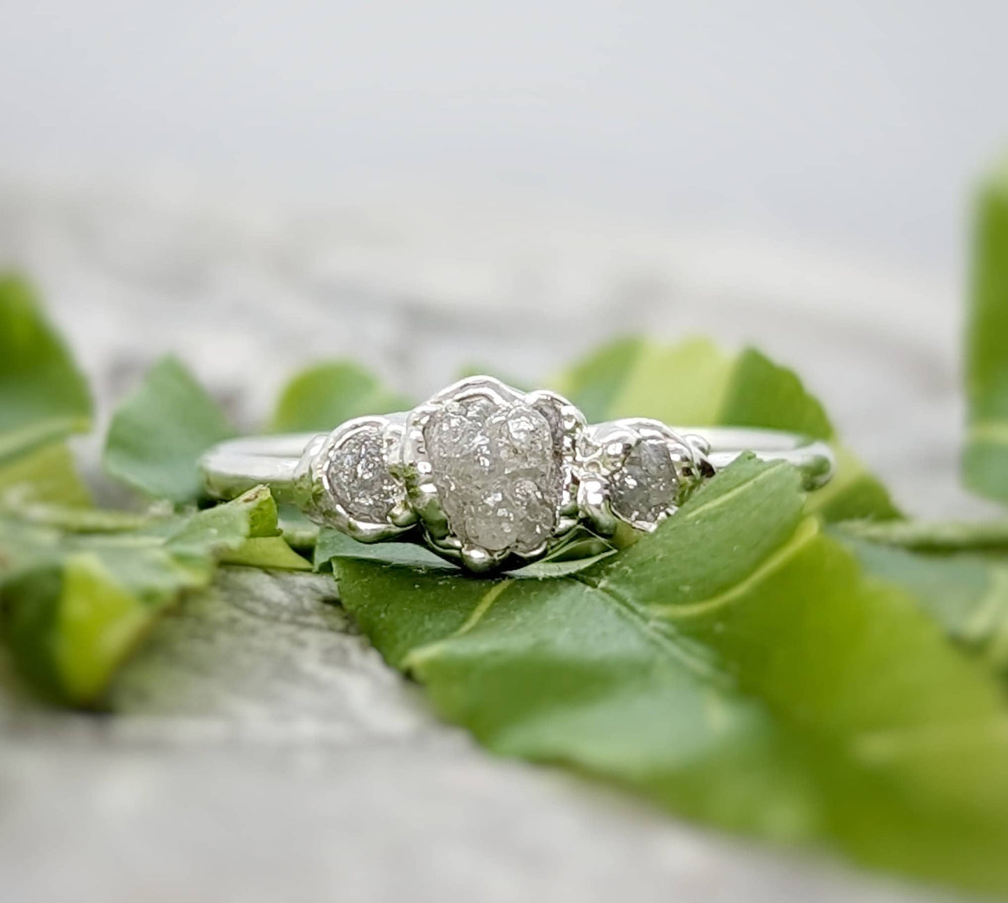 Raw natural diamond engagement ring with a large diamond and two smaller diamonds, surrounded by green leaves.