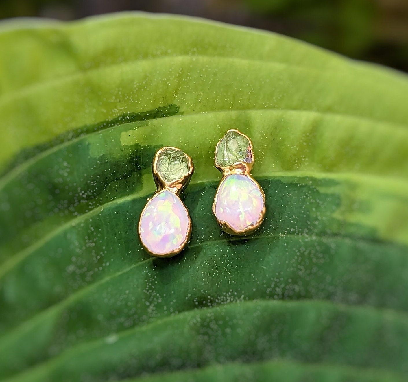 Dainty raw Peridot and Opal stud earrings nestled on green leaves, showcasing unique handcrafted design.
