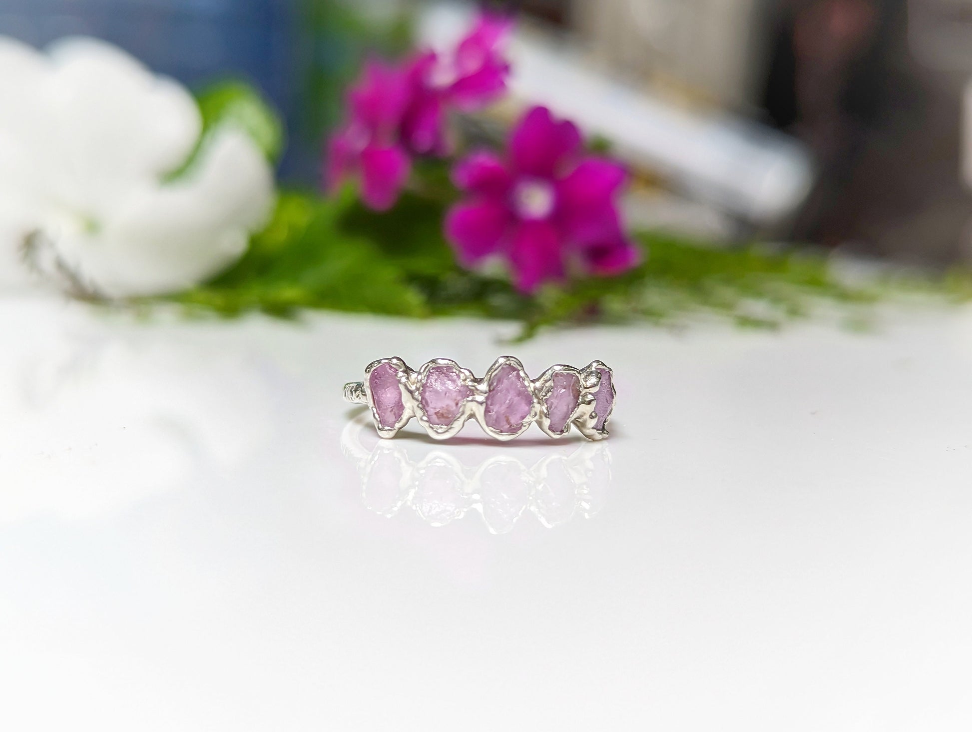Raw pink sapphire eternity ring set on silver band, perfect for gifts or weddings, surrounded by pink flowers.