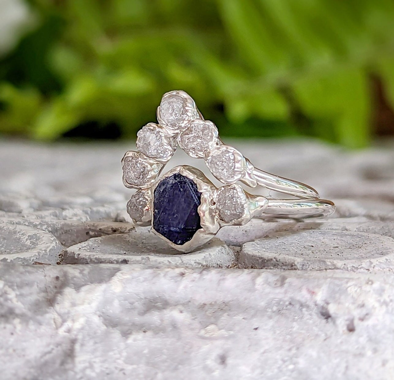Raw sapphire and diamond wedding ring set on a stone surface, featuring a stylish Sterling Silver band.