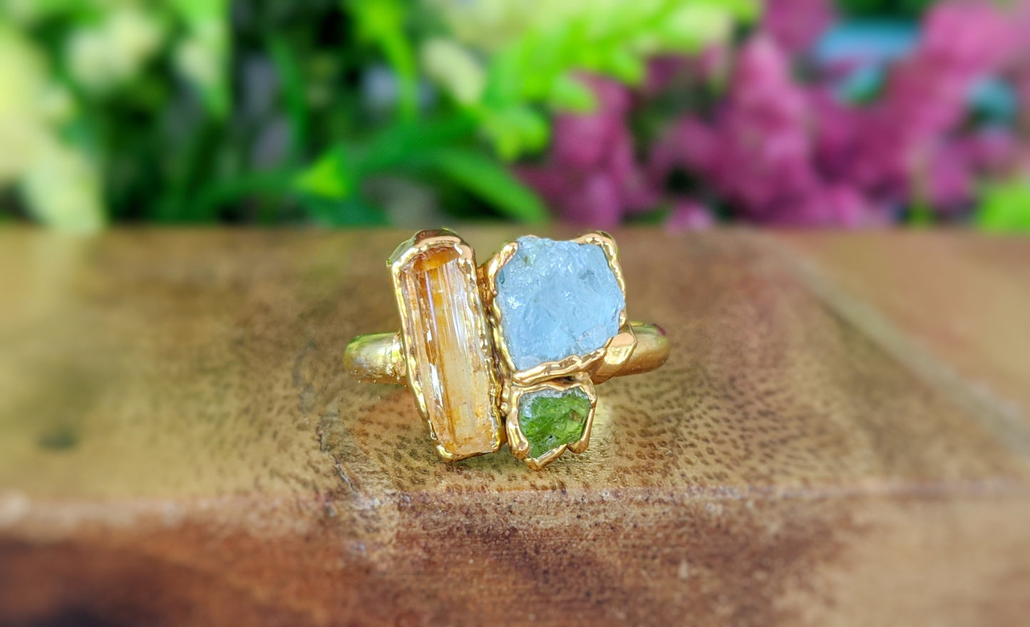 Raw Topaz, Aquamarine and Peridot cluster ring on a wooden surface with a floral background.